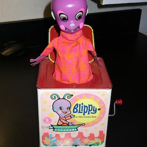 May include: A vintage wind-up toy music box featuring a purple alien character named "Blippy" in a spaceship. The box is red and white with a yellow sun and pink flowers. The text "Blippy in-the-music-box" is printed on the front.