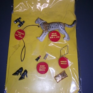 May include: A yellow plastic bag with a variety of items inside, including a toy ocelot with movable head and legs, a pair of binoculars, a lipstick whistle, a handcuff bracelet, and a small purse. The bag has red and white text that reads "Ocelot Movable Head & Legs", "Binoculars with Real Lenses", "Secret Lipstick Whistle", and "Handcuff Bracelet".