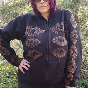 May include: Black zip-up hoodie with a gold eye design printed on the front and sleeves. The hoodie has a hood and a zipper closure.