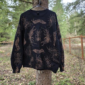 Puede incluir: Sudadera negra con un patrón repetido de insectos en color bronce, incluyendo hormigas y ciempiés. La prenda se muestra al aire libre, colgada de un árbol. El diseño presenta varios artrópodos.