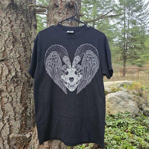Pode incluir: Camiseta preta com um design gráfico branco. O design apresenta um crânio com dois pássaros e asas, formando um formato de coração. A camiseta está pendurada em um cabide preto em um cenário natural ao ar livre.