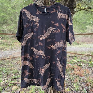 May include: Black t-shirt with a repeating pattern of tan slugs. The shirt has a crew neck and short sleeves. The design appears to be bleached onto the fabric, creating a unique, distressed look. The shirt is hanging on a black hanger.