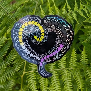 Puede incluir: Una pegatina en forma de corazón con un diseño de ciempiés en los colores de la bandera del orgullo no binario: amarillo, blanco, negro, morado e iridiscente. La pegatina está sobre un fondo de hojas verdes de helecho.