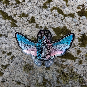 Puede incluir: Una pegatina de un escarabajo con las alas extendidas, las alas son un degradado de azul y rosa. El escarabajo es negro con un diseño detallado.