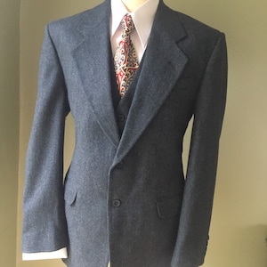 Puede incluir: Un traje de tres piezas gris azulado oscuro, exhibido en un maniquí. El traje incluye una chaqueta, un chaleco y una camisa con corbata estampada. La chaqueta tiene cierre de dos botones y solapas con muescas. Un traje clásico.