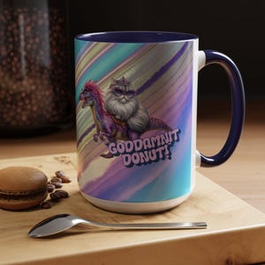 May include: A white ceramic mug with a navy blue interior and handle. The mug features a colorful illustration of a cat riding a dinosaur with the text "GODDAMNIT DONUT!". The background has a colorful, abstract design.