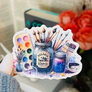 May include: A sticker featuring a watercolor illustration of a jar filled with paintbrushes, a jar of blue liquid with the text "The Velaris Soup Co. Chapter 53", a watercolor palette, and a jar with a galaxy design. The sticker has a white background with a blue, purple, and pink watercolor wash.
