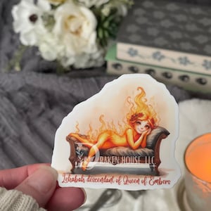 May include: A sticker featuring a woman with fiery orange hair and a body made of flames, lying on a couch. The text "Lehabah descendant of Queen of Embers" is printed below the image.