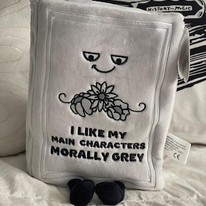 May include: A plush, gray book-shaped pillow with embroidered details. The pillow features a smiling face, a floral design, and the text "I LIKE MY MAIN CHARACTERS MORALLY GREY". Black, rounded feet are at the base.