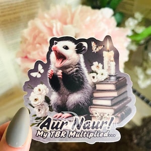May include: A sticker featuring a cartoon opossum with an open mouth, surrounded by books, flowers, and butterflies. The sticker has the text "Aur Nau! My TBR Multiplied..." in a stylized font. The opossum is black, white, and gray.