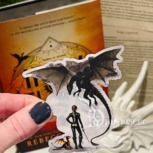 Könnte beinhalten: Ein schwarzer Drache mit ausgebreiteten Flügeln, ein kleiner gelber Drache und eine Person mit einem Schwert und einem dunklen Umhang stehen auf einem weißen Hintergrund. Der Text "DRAKEN HOUSE, LLC." ist in der rechten unteren Ecke sichtbar.