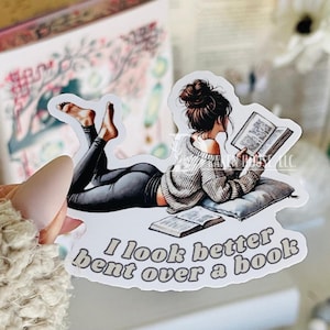 May include: A sticker featuring a woman in a grey sweater and black leggings reading a book. The sticker has the text "I look better bent over a book."