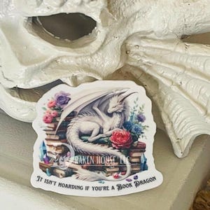 May include: White dragon curled up on a stack of books with flowers and gems. The text reads "It isn't hoarding if you're a book dragon."