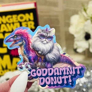 Könnte beinhalten: Ein Aufkleber mit einem Dinosaurier mit rosa und lila Federn und einer Katze mit Sonnenbrille und Krone. Der Aufkleber hat den Text "GODDAMNIT DONUT!" in Pink und Blau und den Text "RAKEN HOUSE LLC".