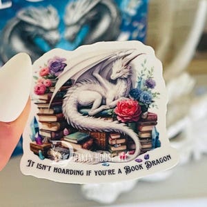May include: A white dragon with wings curled around a stack of books. The dragon is surrounded by flowers and crystals. The text "It isn't hoarding if you're a book dragon" is at the bottom of the image.