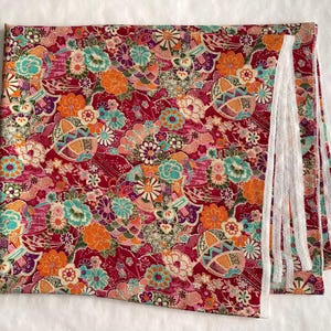 May include: A rectangular piece of fabric featuring a vibrant red background with a dense, multicolored floral pattern including orange, teal, purple, and white blossoms, along with geometric fan shapes and white lace trim along one edge.