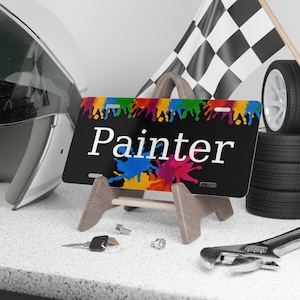 May include: A black license plate with colorful paint splatters and the word "Painter" in white text. The plate is on a wooden easel.