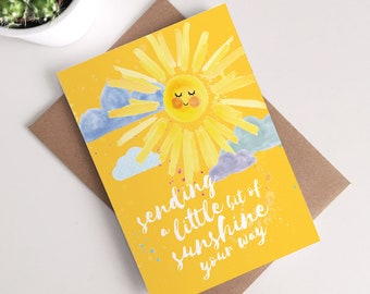 May include: A yellow greeting card with a smiling sun illustration and the text "sending a little bit of sunshine your way." The card is on a brown envelope, with a small potted plant in the background.