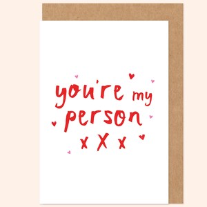 Puede incluir: Una tarjeta de felicitación con fondo blanco y borde marrón. La tarjeta presenta el texto rojo "you're my person" con tres símbolos "X" rojos e ilustraciones de pequeños corazones y puntos.