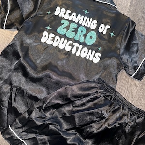 May include: Black satin pajama set with white trim. The top features short sleeves and a collar. The text "Dreaming of Zero Deductions" is printed on the back in white and teal glitter. The set includes a matching pair of shorts.
