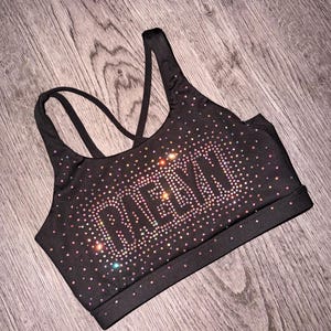 May include: Black sports bra with iridescent rhinestone embellishments spelling out a word. The bra has a racerback design with thin straps and a wide band at the bottom. The rhinestones shimmer in various colors.