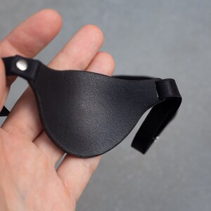 May include: A black leather eye patch with an adjustable elastic strap. The patch is held in a hand.
