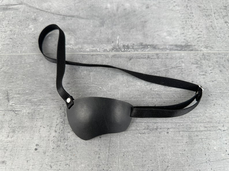 May include: A black leather eye patch with an adjustable strap. The patch is designed to cover one eye and has a simple, classic style.