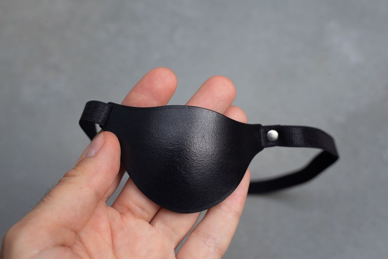 May include: A black leather eye patch with an adjustable strap. The patch is held in a hand.