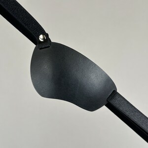 May include: A black eye patch with a soft, padded surface and an adjustable strap. The strap has a metal buckle for fastening.