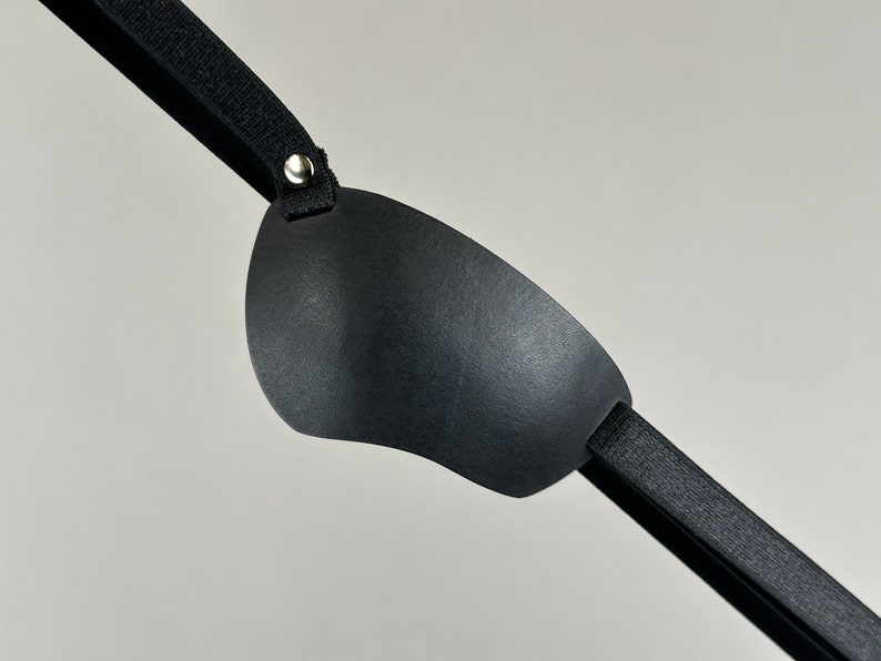May include: A black eye patch with a soft, padded surface and an adjustable strap. The strap has a metal buckle for fastening.