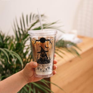 May include: A clear plastic tumbler with a lid and straw, filled with an iced beverage. The exterior features a black graphic print of a person in a witch hat reading a book, with celestial motifs and the text THE READER printed below.
