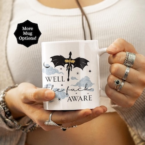 Pode incluir: Caneca de cerâmica branca com um gráfico de dragão preto e o texto "WELL the fuck AWARE". A caneca também apresenta um gráfico preto em forma de estrela que diz "More Mug Options!"