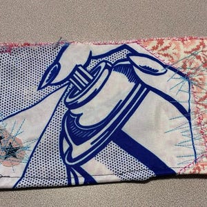 May include: Rectangular fabric patch with a pop art design featuring a blue and white image of a hand holding a spray can. The patch has a pink stitched border and a floral pattern on the right side.