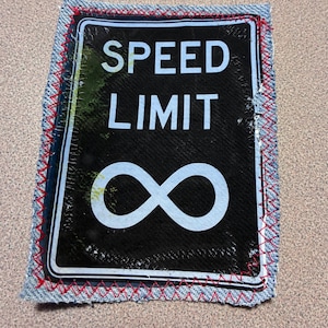 May include: A black sign with the words "SPEED LIMIT" and an infinity symbol in white. The sign is framed with denim fabric and red stitching, creating a unique, handcrafted look. The sign is a creative piece of art.