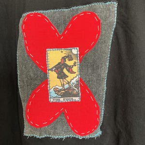 May include: A black t-shirt featuring a rectangular gray denim patch with a bright red fabric cross stitched on top. Centered on the cross is a small rectangular tarot card print titled THE FOOL, depicting a figure standing on a cliff edge.