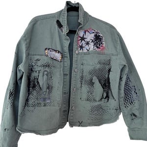 May include: An olive green denim jacket featuring a raw hem, a button-up front, and various embellishments including black mesh-pattern screen prints, a large safety pin graphic, a patch with black stars, and metallic studs on the chest pocket.