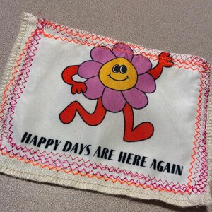 May include: A rectangular patch with a cartoon flower character in pink, yellow, and red, with the text "HAPPY DAYS ARE HERE AGAIN" in black. The patch has a white background with orange and pink stitching.
