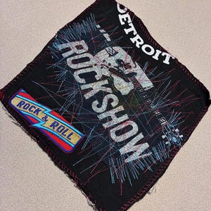 May include: Black square fabric with embroidered text and graphics. The words "ROCK SHOW" and "DETROIT" are visible. A smaller patch reads "ROCK & ROLL" with a lightning bolt design. Red and blue stitching accents the design.