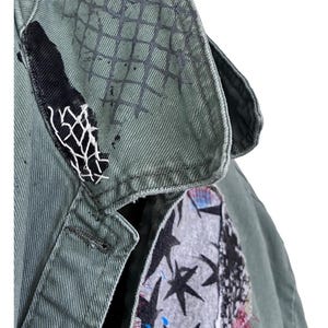 May include: An olive green jacket featuring a collar with a black grid pattern, a black fabric patch with white stitching, and a chest panel displaying a grey fabric insert decorated with black stars and small blue paint splatters.