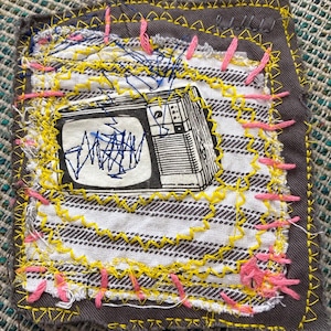 May include: Embroidered patch featuring a vintage television illustration in black and white. The patch is stitched onto a gray fabric with yellow and pink thread accents. The background fabric has a striped pattern.