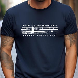 May include: Navy blue t-shirt with white text that reads "NAVAL SUBMARINE BASE" above a graphic and "GROTON, CONNECTICUT". The shirt is a crew neck style.
