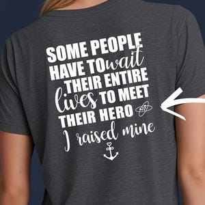May include: A dark gray short-sleeved t-shirt featuring white text on the back that reads: SOME PEOPLE HAVE TO WAIT THEIR ENTIRE LIVES TO MEET THEIR HERO I raised mine, accompanied by a small atom icon and an anchor symbol with a heart at the top.
