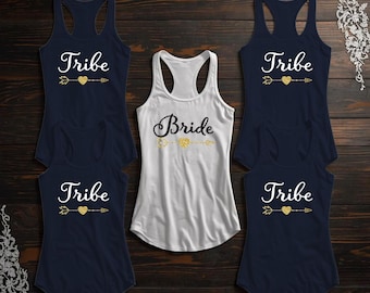 May include: A collection of navy blue and white racerback tank tops. The navy tops say "Tribe" with a gold heart and arrow design. The white tank top says "Bride" in black script with a gold heart and arrow design.