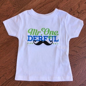 May include: White short-sleeved t-shirt with green and blue text that reads "Mr. One Derful" and a black mustache graphic.