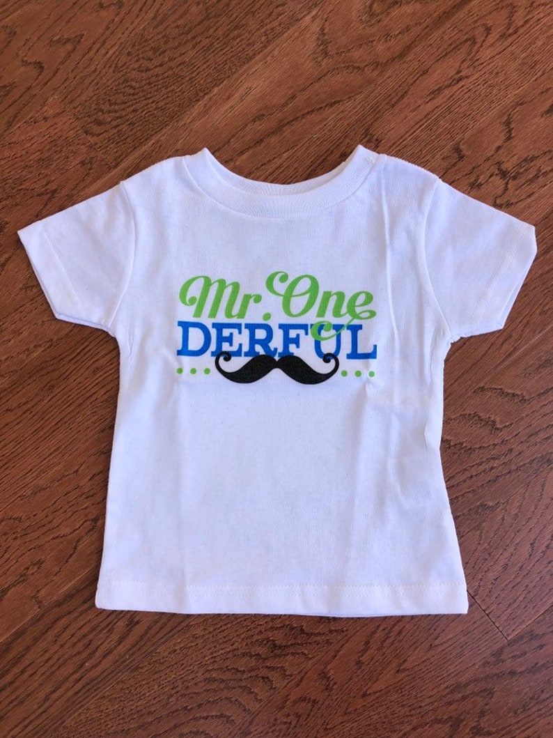 May include: White short-sleeved t-shirt with green and blue text that reads "Mr. One Derful" and a black mustache graphic.