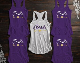 May include: A collection of tank tops, perfect for a bridal party. The shirts are purple and white, with the word "Tribe" printed on the purple shirts and "Bride" on the white shirt. The text is accompanied by a gold heart and arrow design.