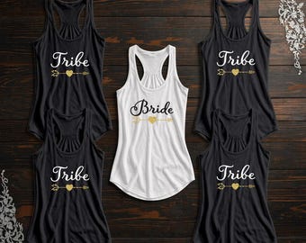 May include: A collection of black and white racerback tank tops. The black tank tops have the word "Tribe" printed in white with a gold arrow and heart design. The white tank top has the word "Bride" in black with a gold arrow and heart design.