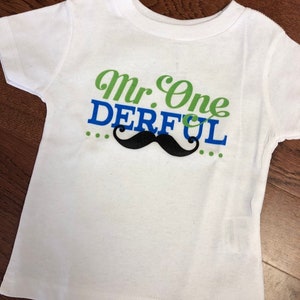 May include: White short-sleeved t-shirt with green and blue text that reads "Mr. One Derful" with a black mustache graphic.