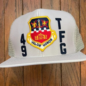 May include: A light gray trucker hat with a mesh back and a curved brim. The front features a patch with a shield design in red, yellow, and black, with the words "VALOR HONOR". The numbers "49" and letters "TFG" are also displayed.