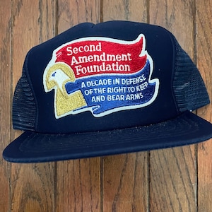 Op de afbeelding: Een marineblauwe truckerpet met een mesh achterkant en een gebogen klep. De voorkant heeft een patch met de tekst "Second Amendment Foundation" in rood en wit, met blauwe tekst en een gouden embleem.